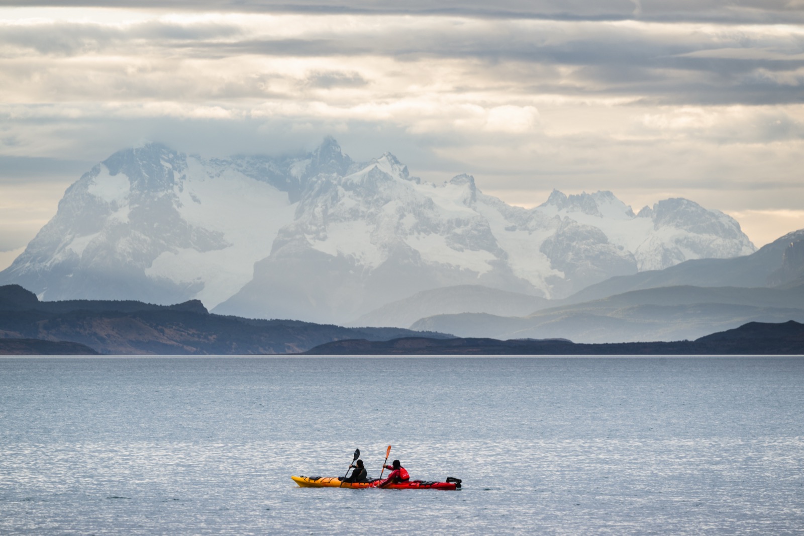 Two people kayaking on calm water toward snow-capped Patagonian peaks