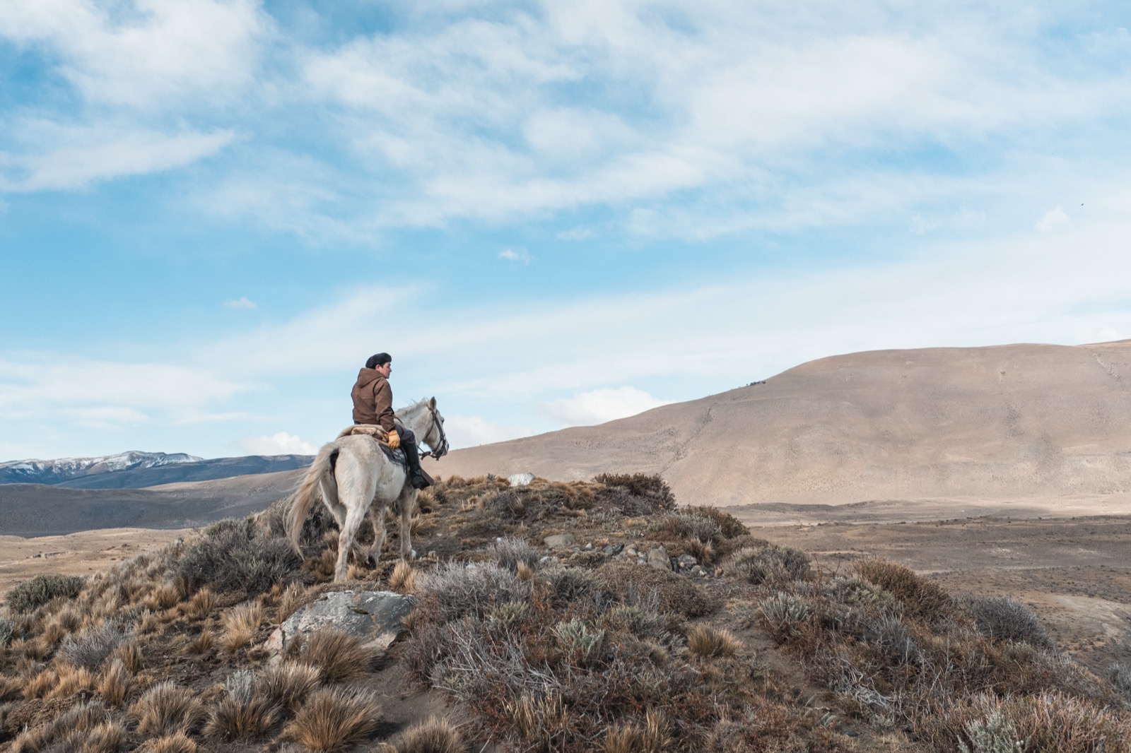Two riders on horseback crossing the golden Patagonian steppe by a lake, snow-dusted mountains behind